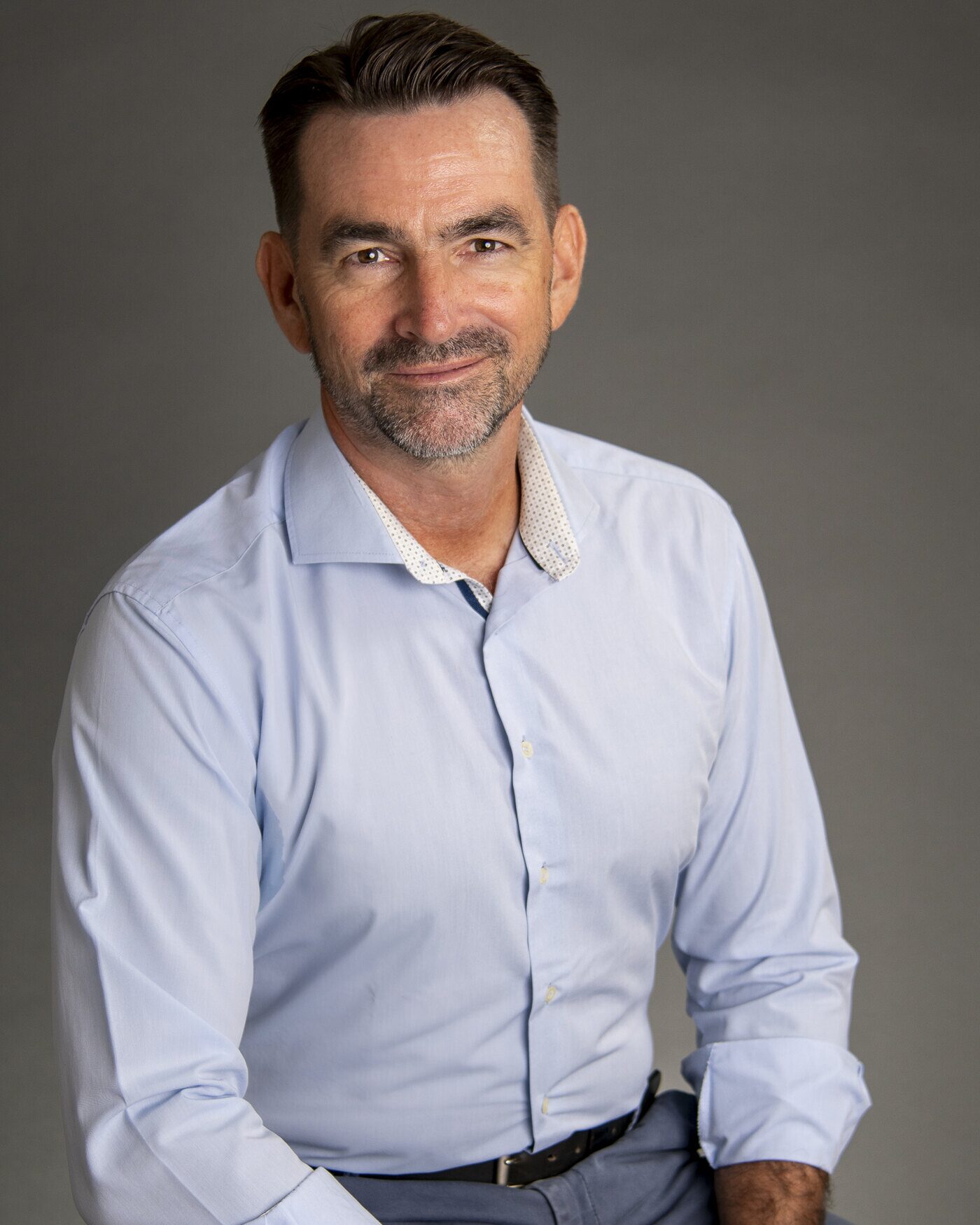 Morgan Meinecke, seated, in a light blue shirt against a grey studio backdrop