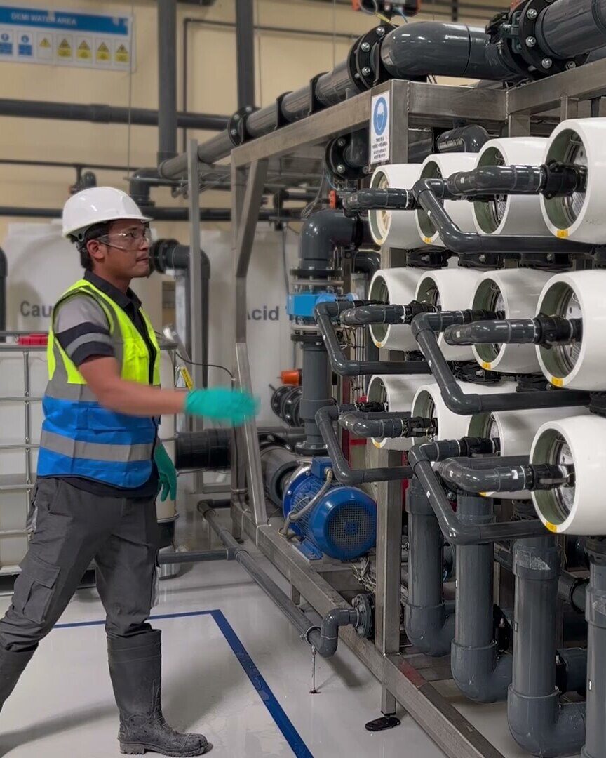 An operator in hard hat, glasses, hi-vis vest and gloves at the regeneration rig in the Second Life RO plant, pressure vessels piped up beside him
